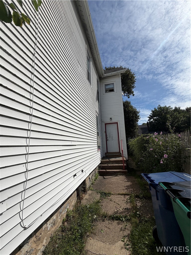 White-sided house with a narrow path to a side entrance, pink flowers, and bins.