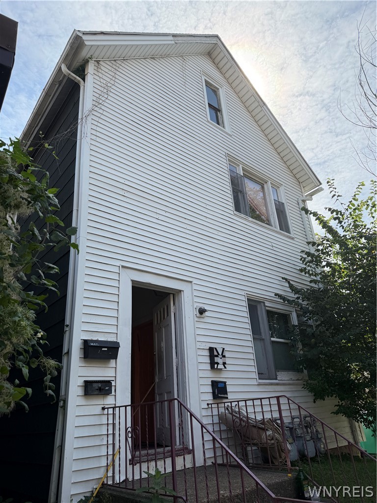 Tall white-sided house with a steep gabled roof and open front entrance above metal-railed steps.