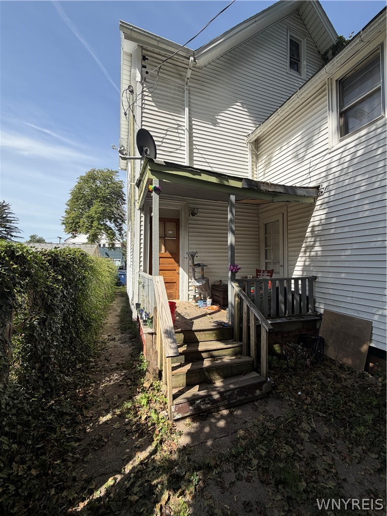White-sided house with a wooden side porch and steps beside a tall hedge.