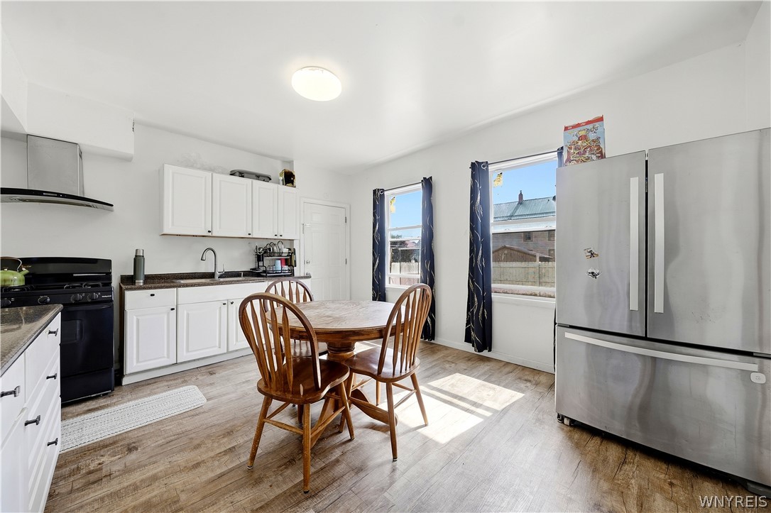Kitchen with white cabinets, stainless steel refrigerator, and a wooden dining table beside sunny windows.