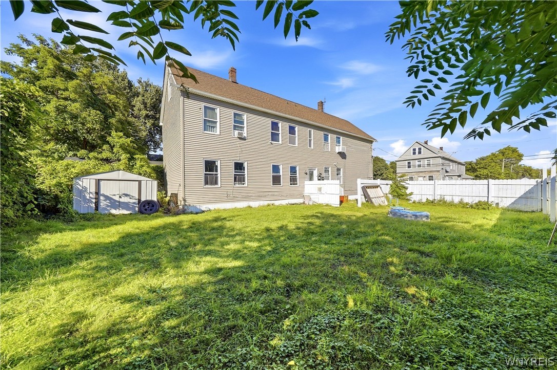 Two-story beige house behind a grassy backyard with a white fence and small shed.