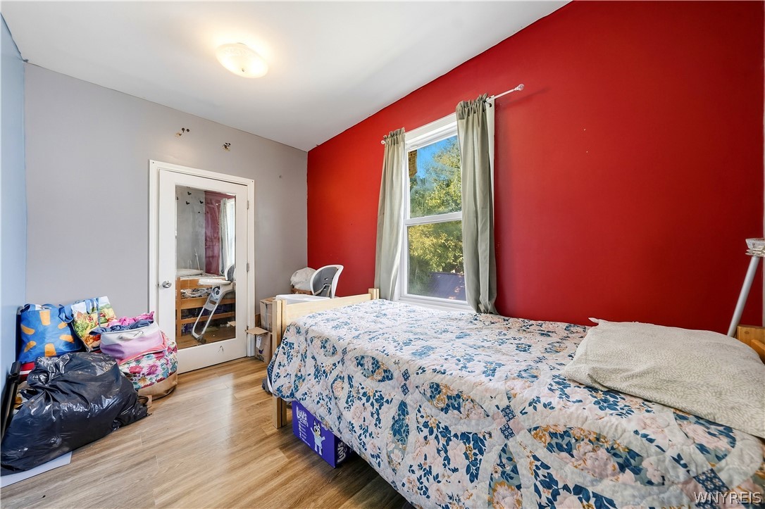 Bedroom with a floral quilt, bold red accent wall, and belongings piled beside the door.