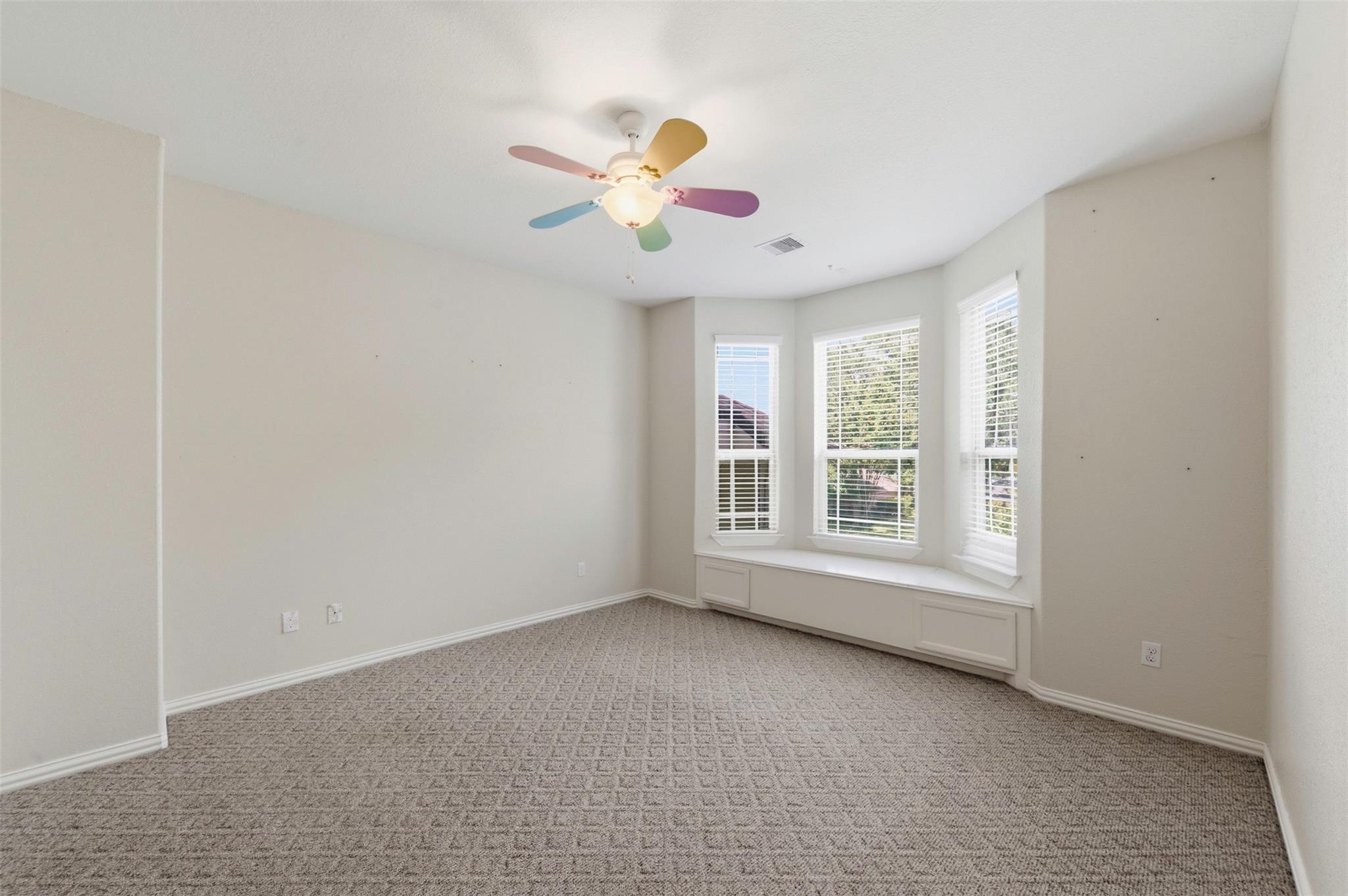 Empty bedroom with a carpeted floor, bay-window seat, and colorful ceiling fan.