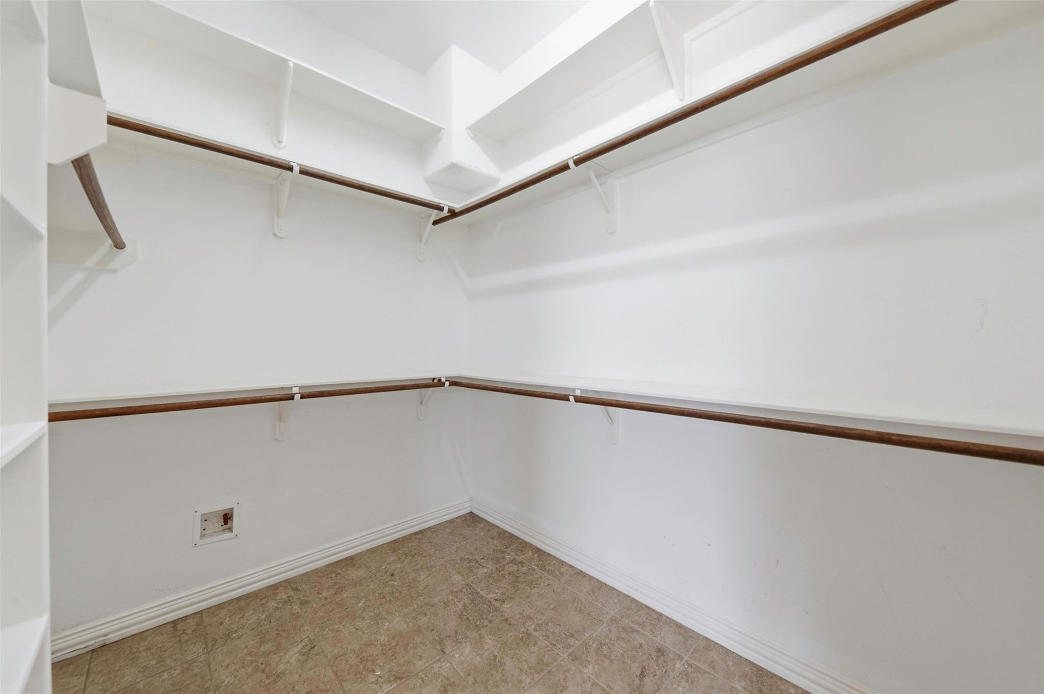 Empty walk-in closet with white shelves, dark wood hanging rods, and tan tile flooring.