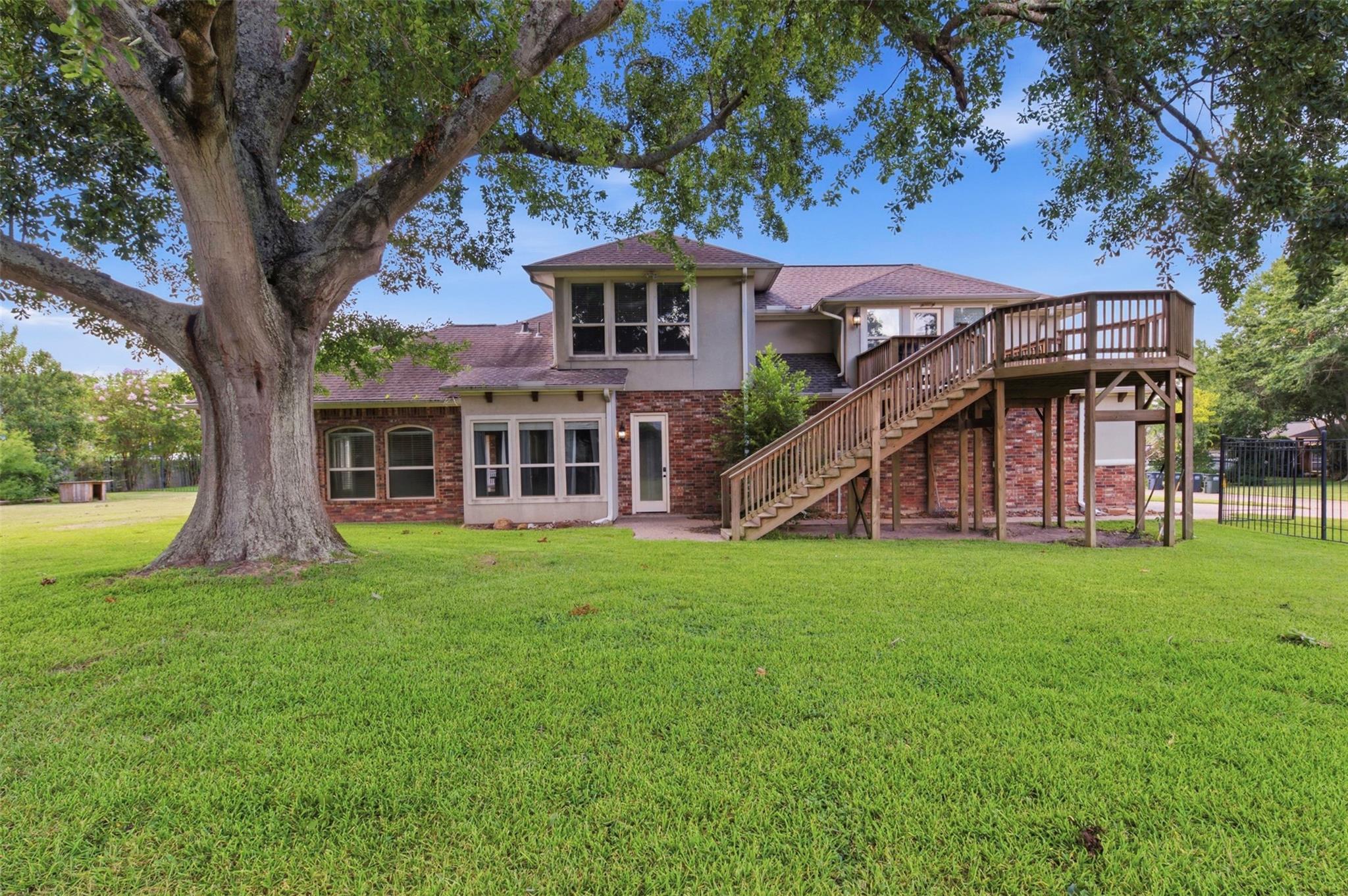 Rear of a two-story brick house with a raised wooden deck and staircase, lawn, and large shade tree.