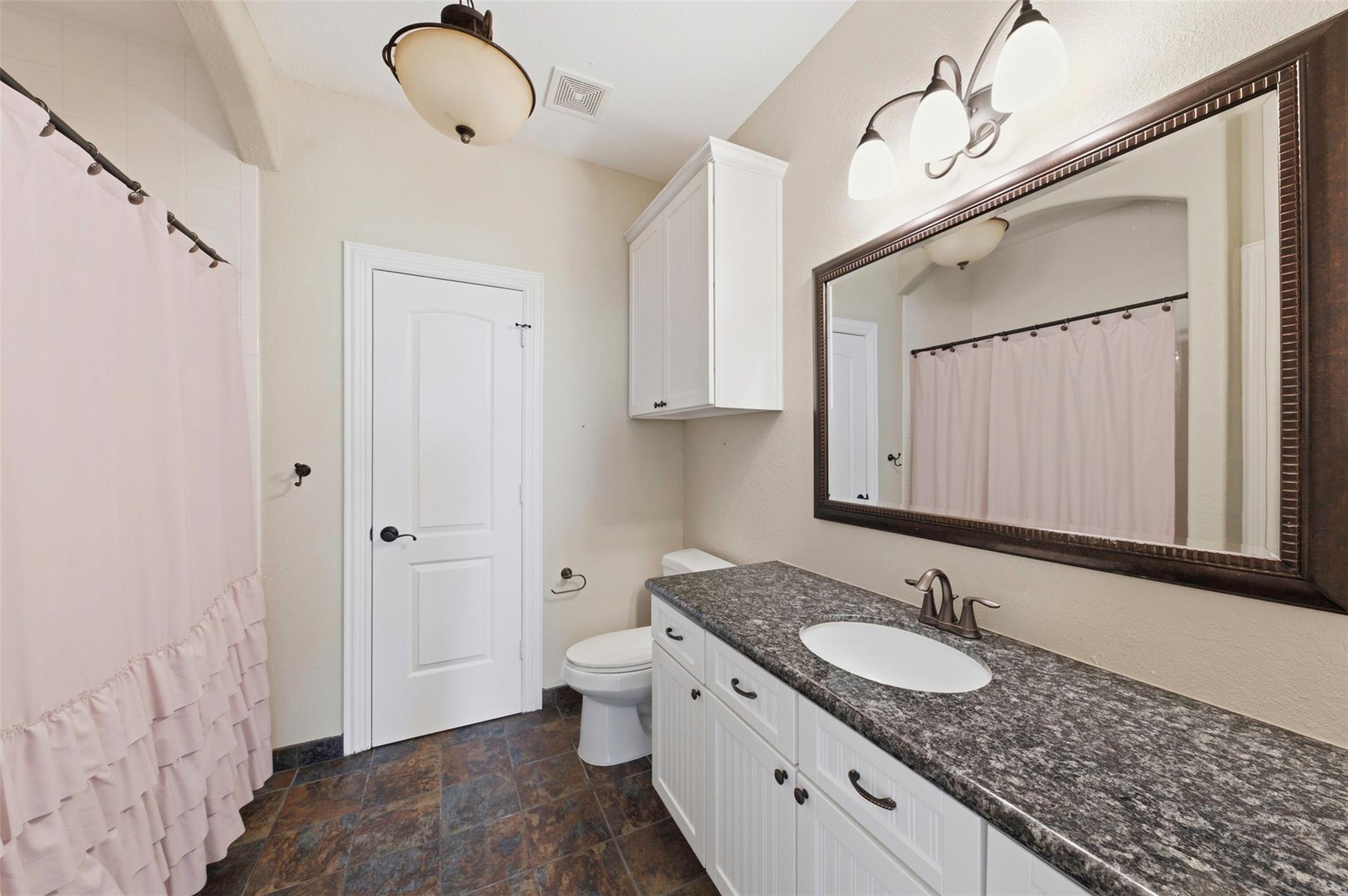 Bathroom with a long granite-topped vanity, large framed mirror, and pink ruffled shower curtain.