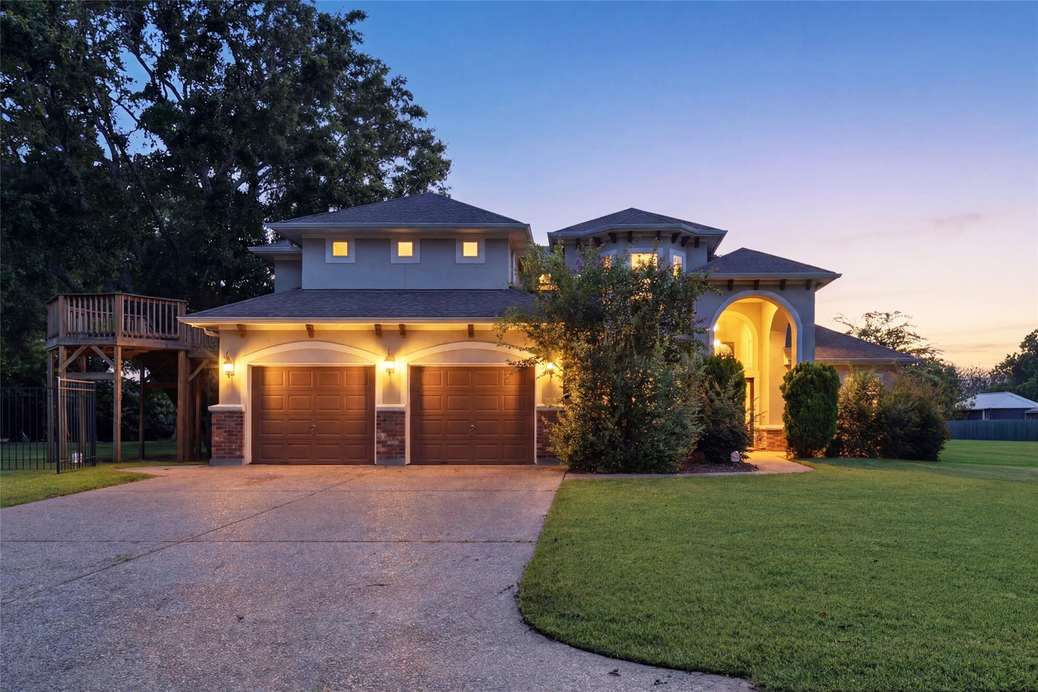Two-story gray home with a double garage, illuminated arched entry, and raised wooden deck at dusk.