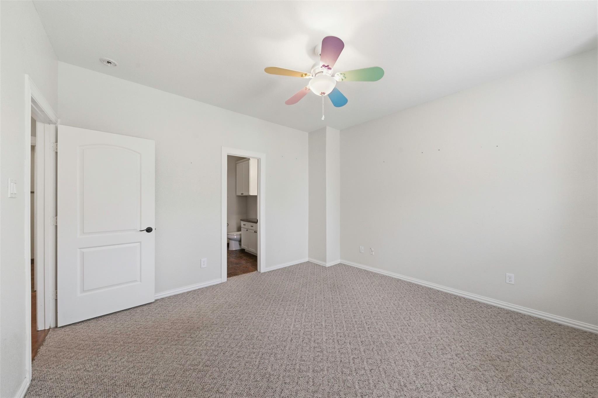 Empty carpeted bedroom with a colorful ceiling fan and an adjoining bathroom doorway.
