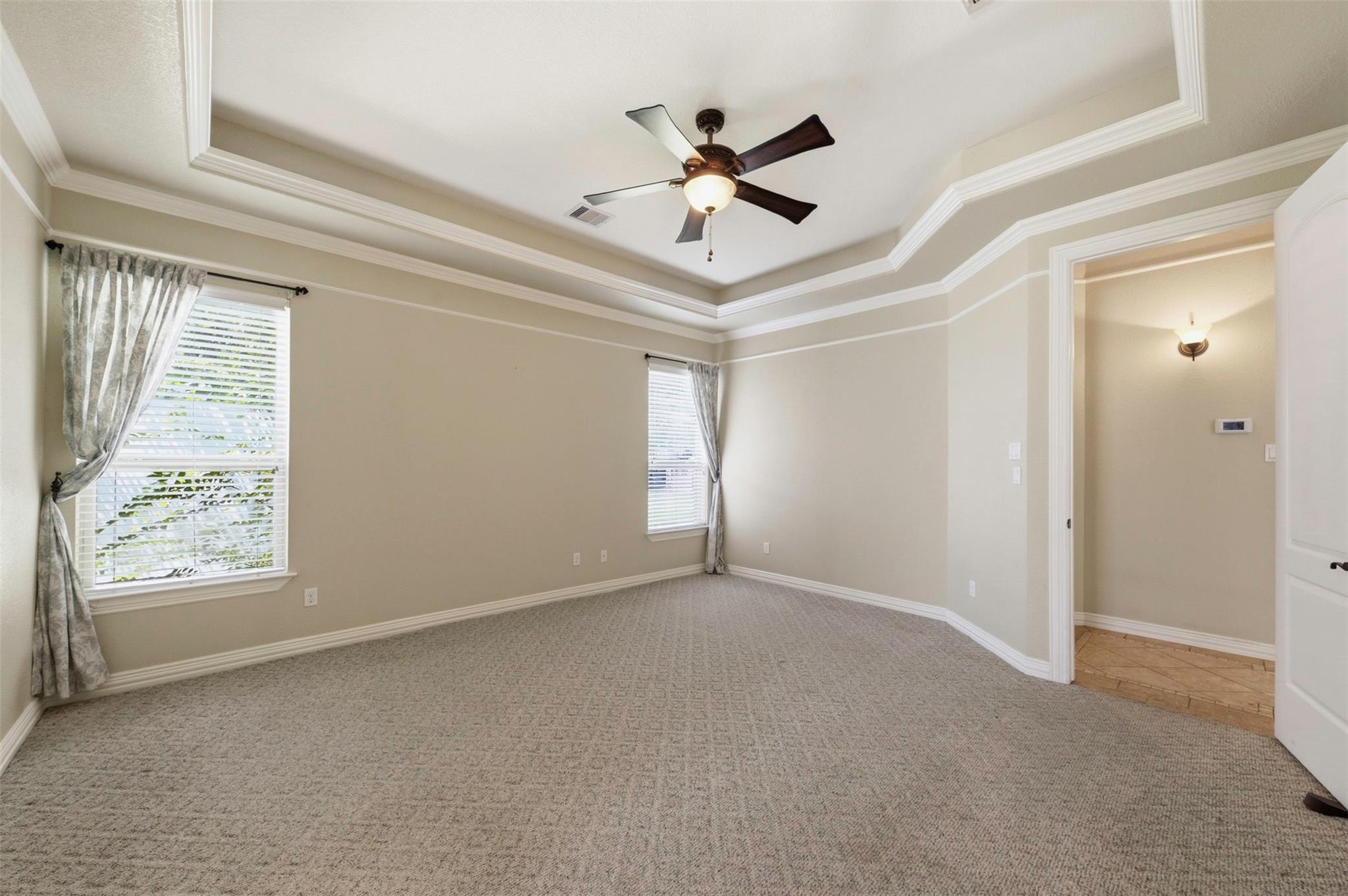 Empty carpeted bedroom with a tray ceiling, ceiling fan, and two curtained windows.