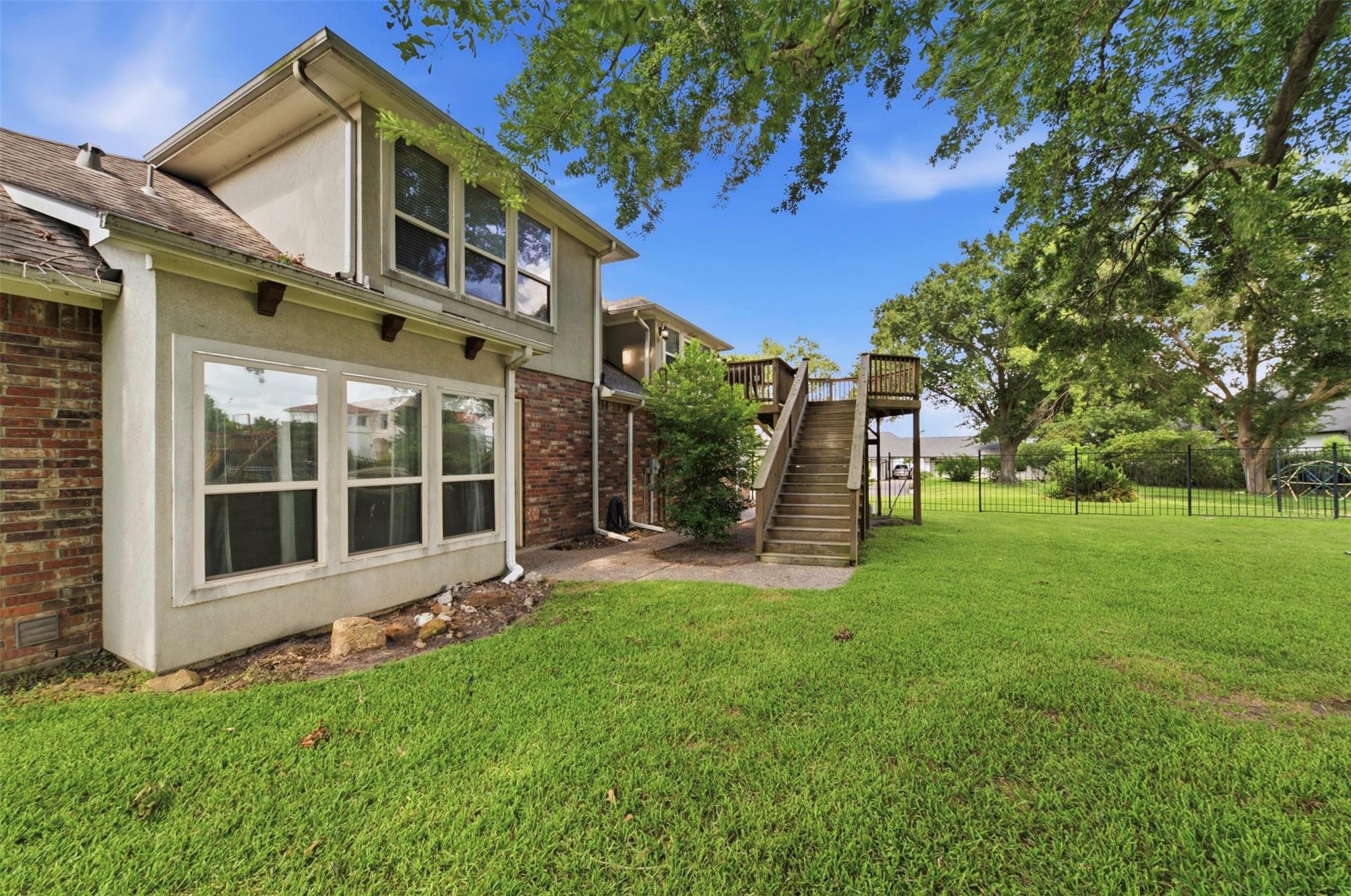 Two-story home’s backyard with raised wooden deck, stairs, fenced lawn, and mature trees.