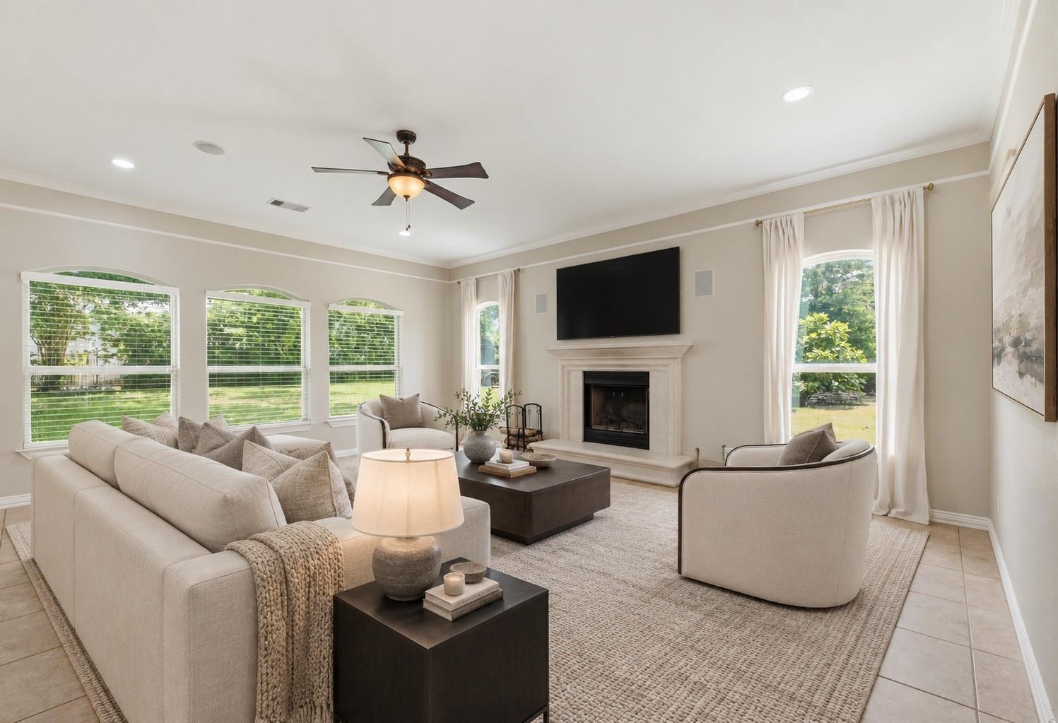 Staged living room with a fireplace, wall-mounted television, and large windows overlooking a green yard.