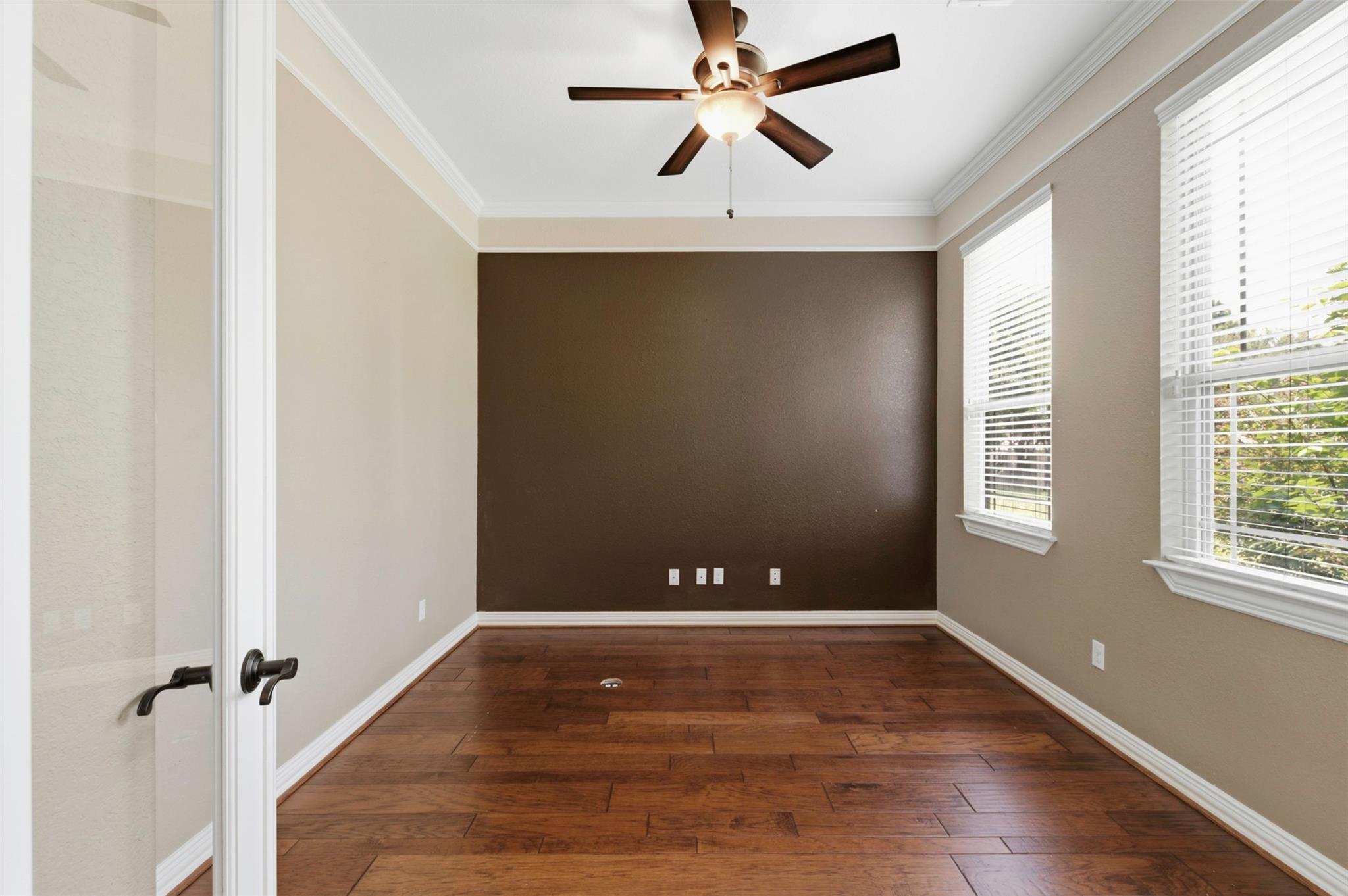 Empty room with a brown accent wall, dark wood floors, two windows, and a ceiling fan.