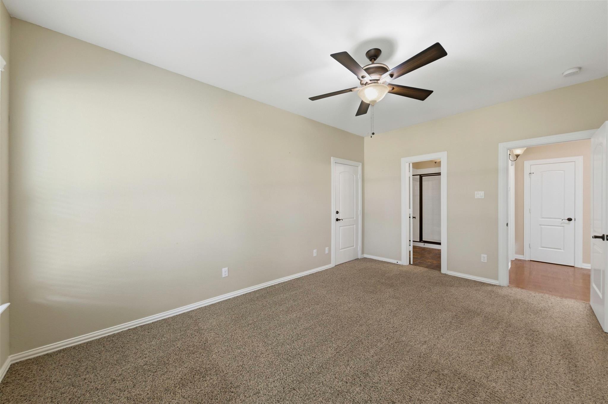 Empty beige bedroom with carpet, a ceiling fan, and open doorways to a closet and hall.