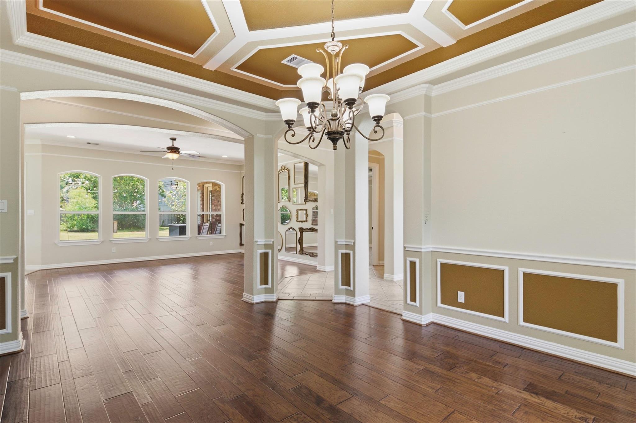 Empty formal dining room with a coffered ceiling, chandelier, and arched windows overlooking greenery.