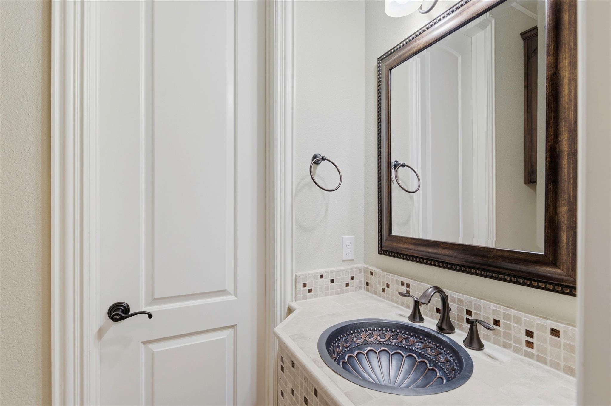 Bathroom vanity with an ornate metal vessel sink, mosaic tile backsplash, and dark framed mirror.