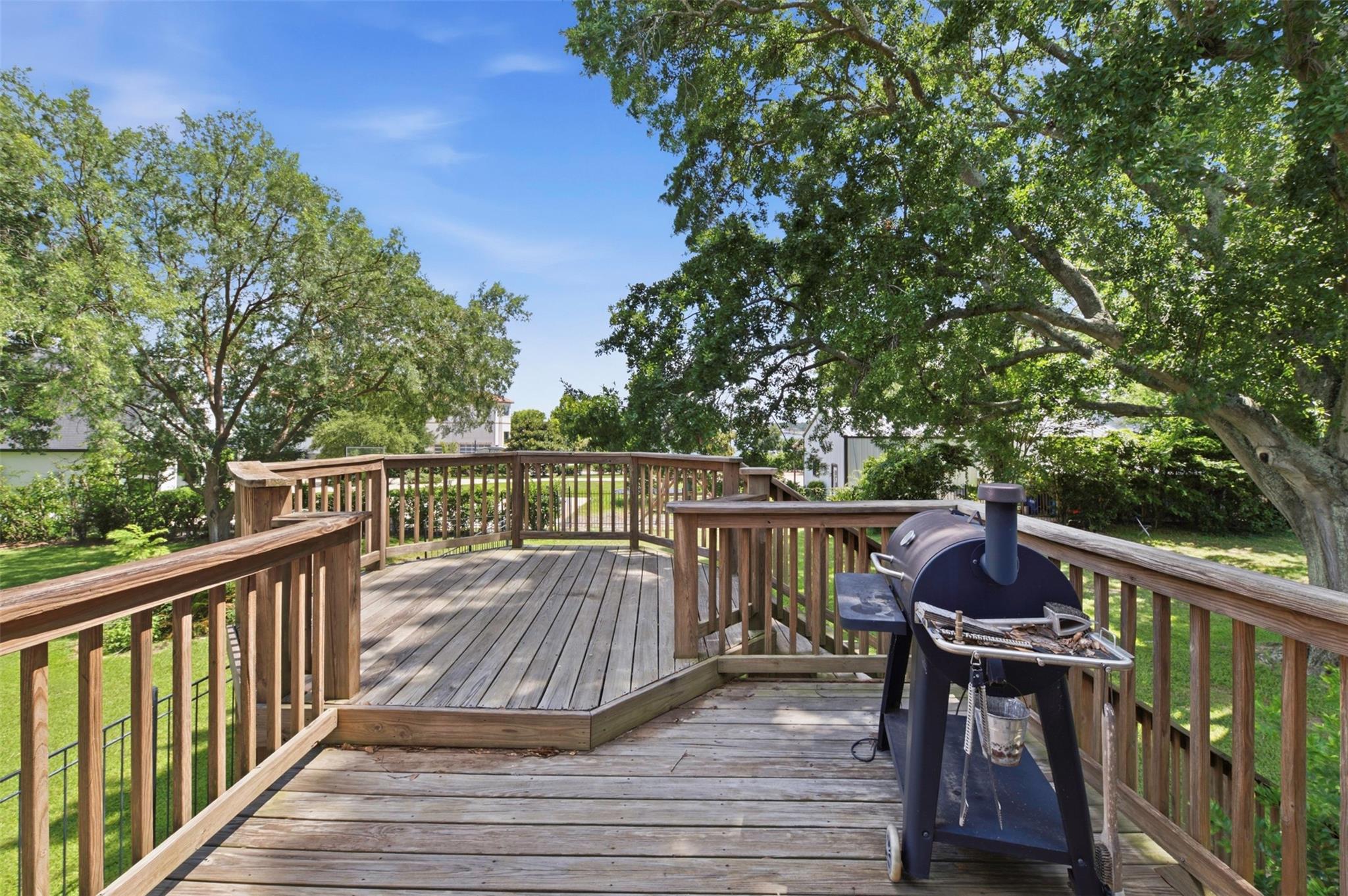 Wooden backyard deck with a smoker grill beneath mature leafy trees.