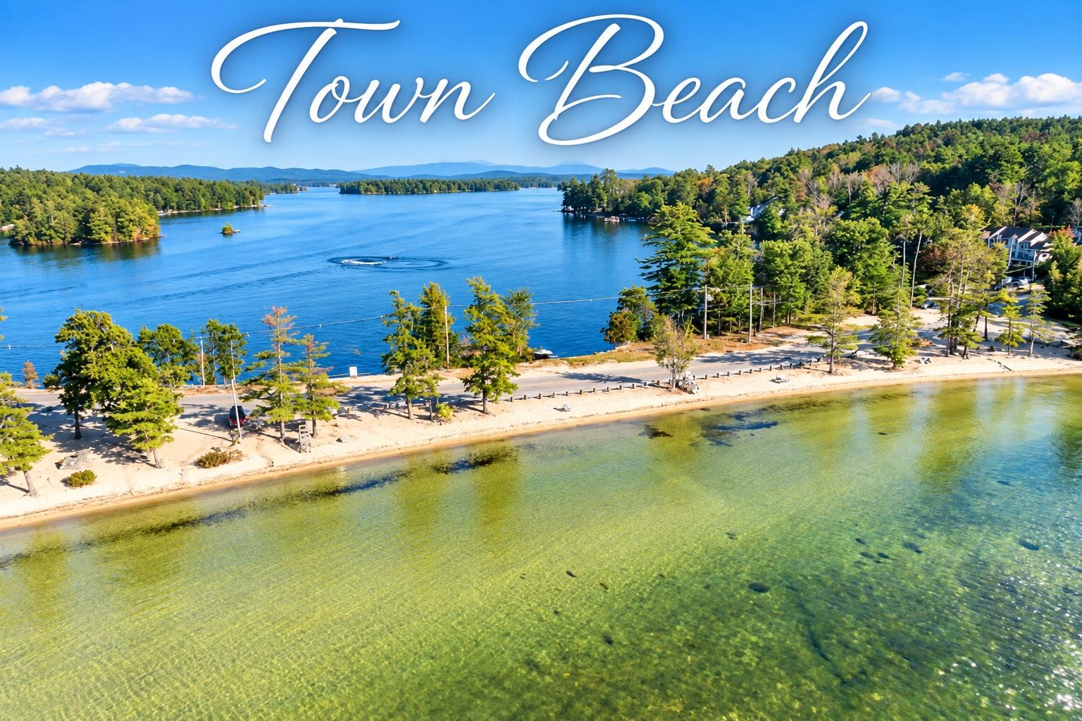 Town beach on a clear lake, with a sandy shoreline, wooded shores, and distant mountains.
