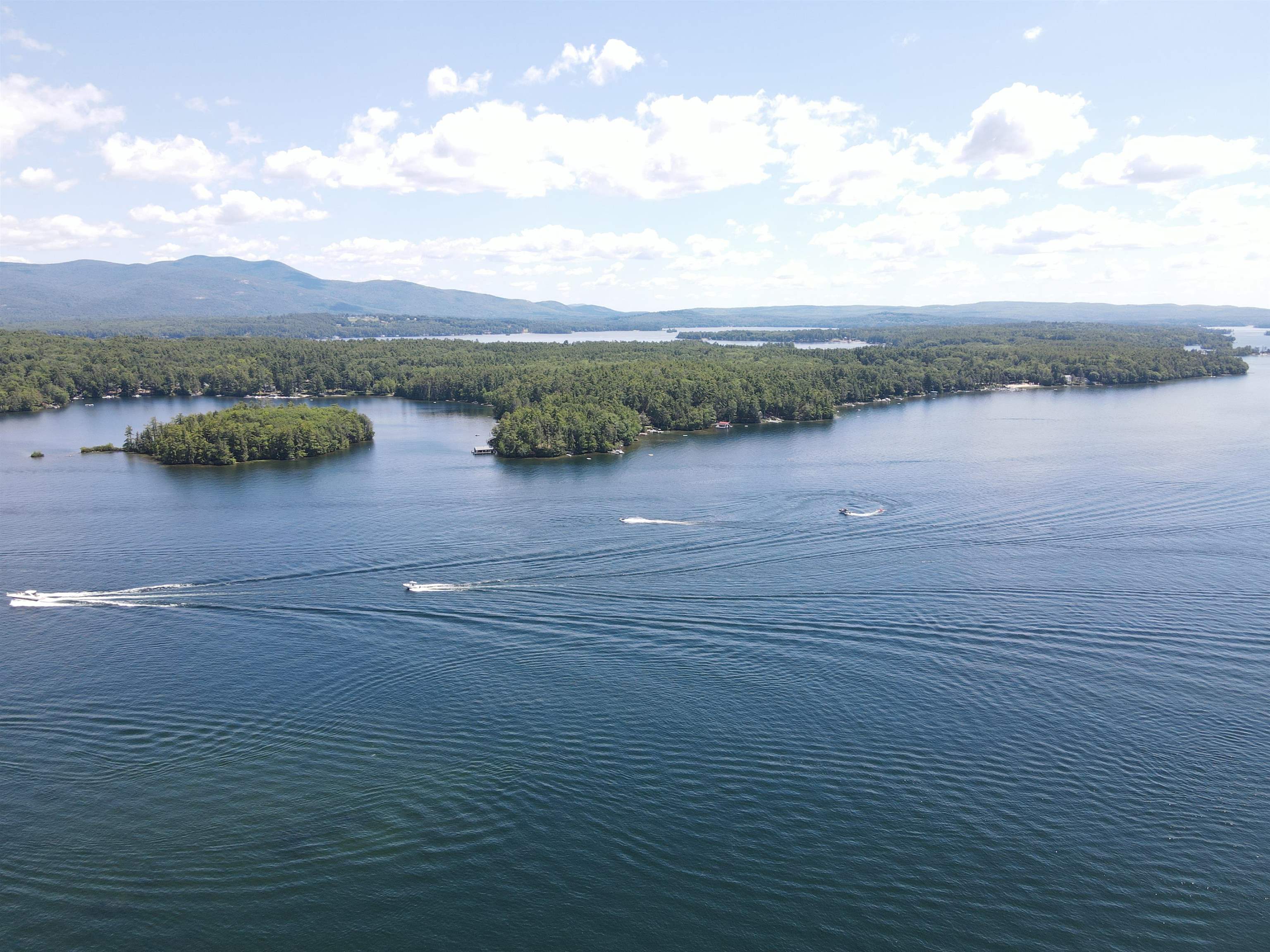 Aerial lake view with wooded islands, boats, and distant mountains.