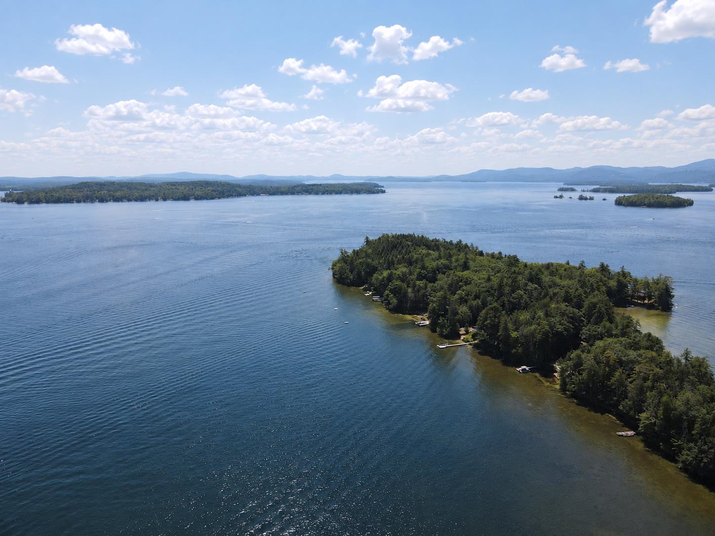 Aerial view over a wooded lakeside peninsula, open water, islands, and distant mountains.