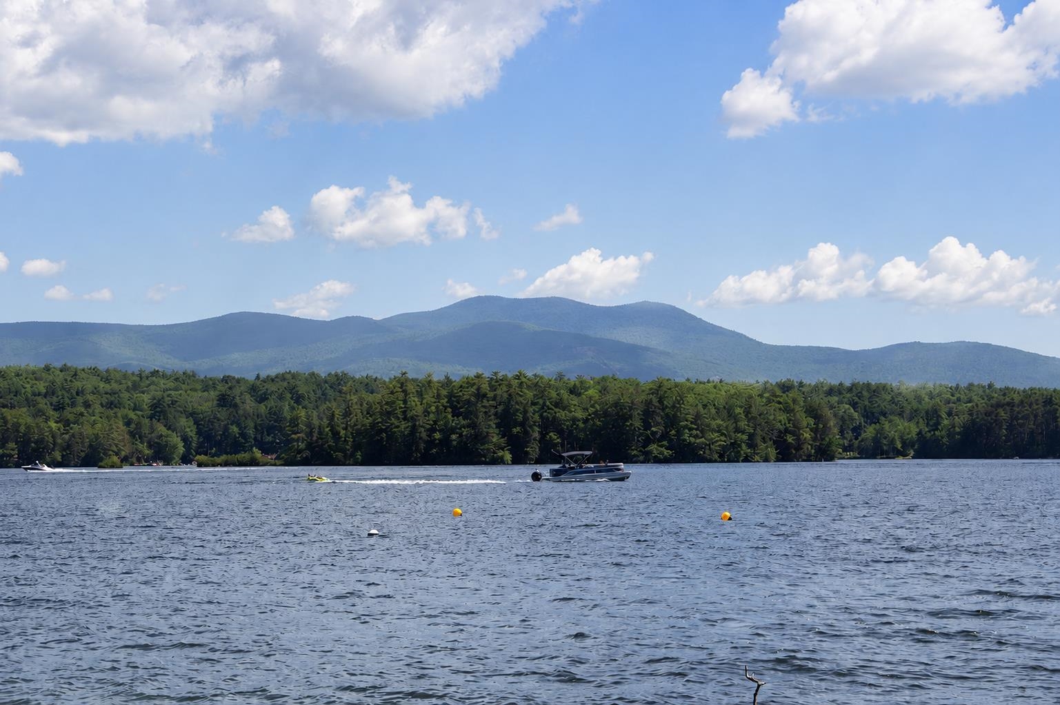 Lake with boats and buoys, backed by a forested shore and distant mountains.