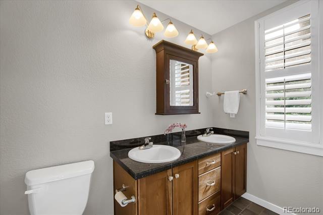 Bathroom with a double-sink vanity, dark countertop, warm wood cabinets, and white plantation shutters.