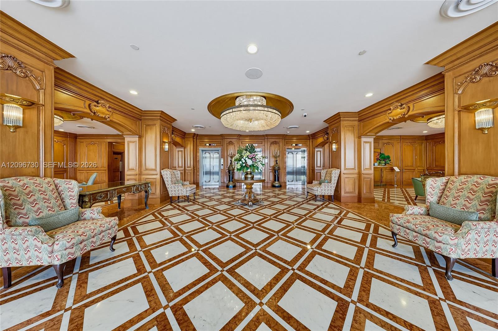 Ornate condominium lobby with carved wood paneling, crystal chandelier, geometric marble floor, and seating.