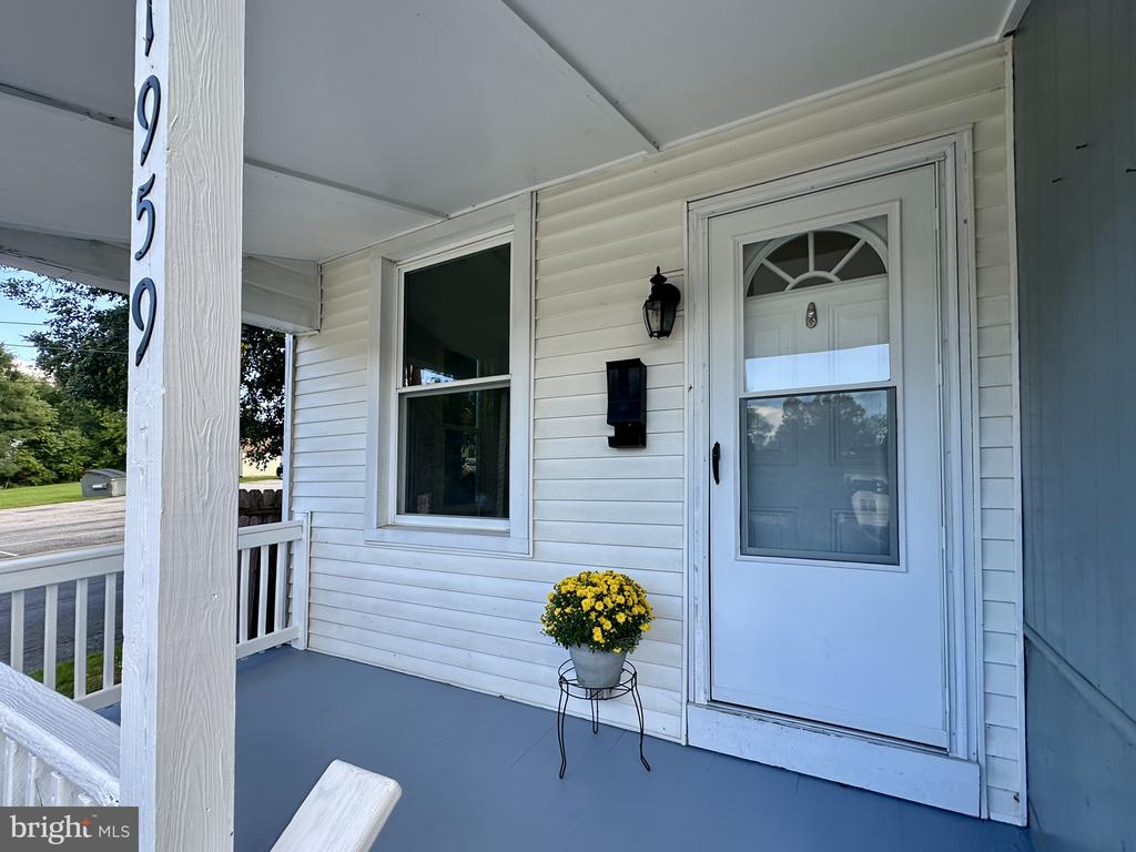 Covered front porch with a white storm door, gray flooring, and yellow flowers beside the entrance.