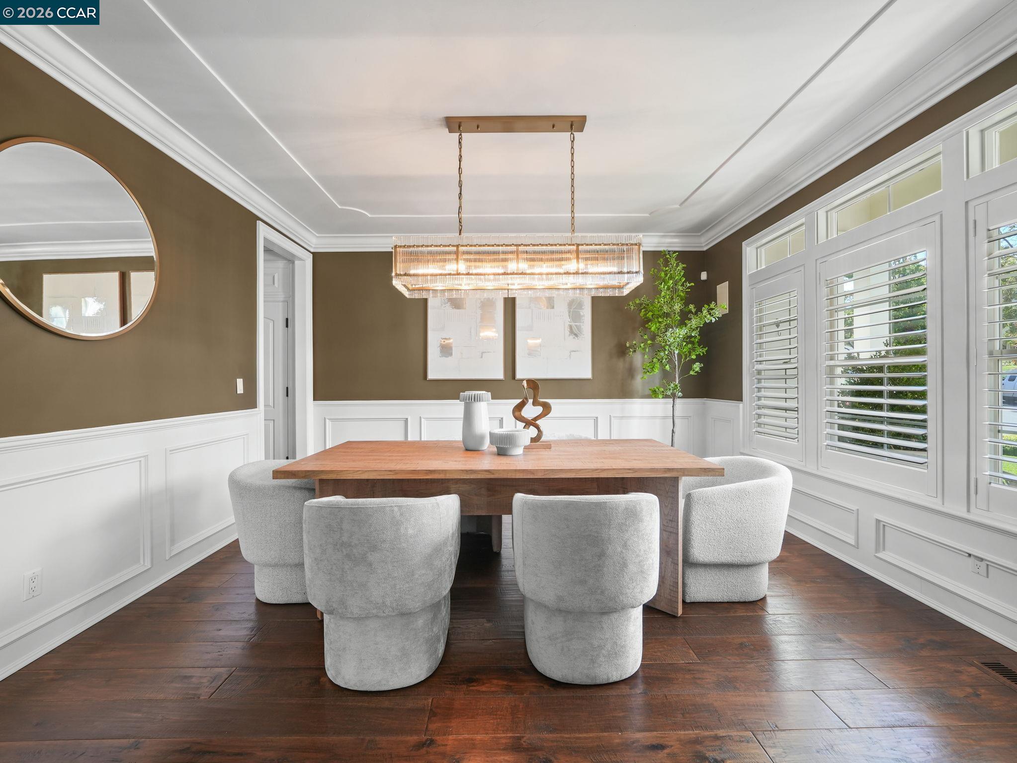Dining room with a wood table, upholstered chairs, linear chandelier, and plantation shutters.