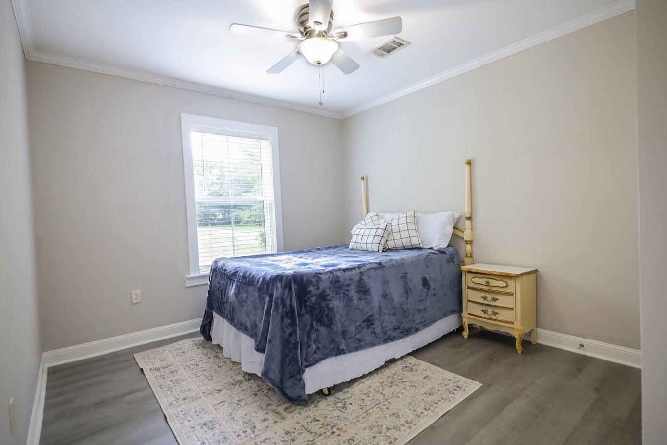 Bedroom with a blue-covered bed, window, ceiling fan, and wooden nightstand.