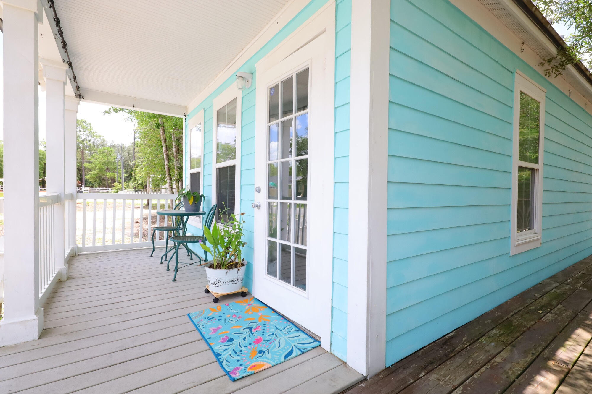 Covered porch beside a turquoise house, furnished with a small table, chairs, potted plants, and a floral doormat.