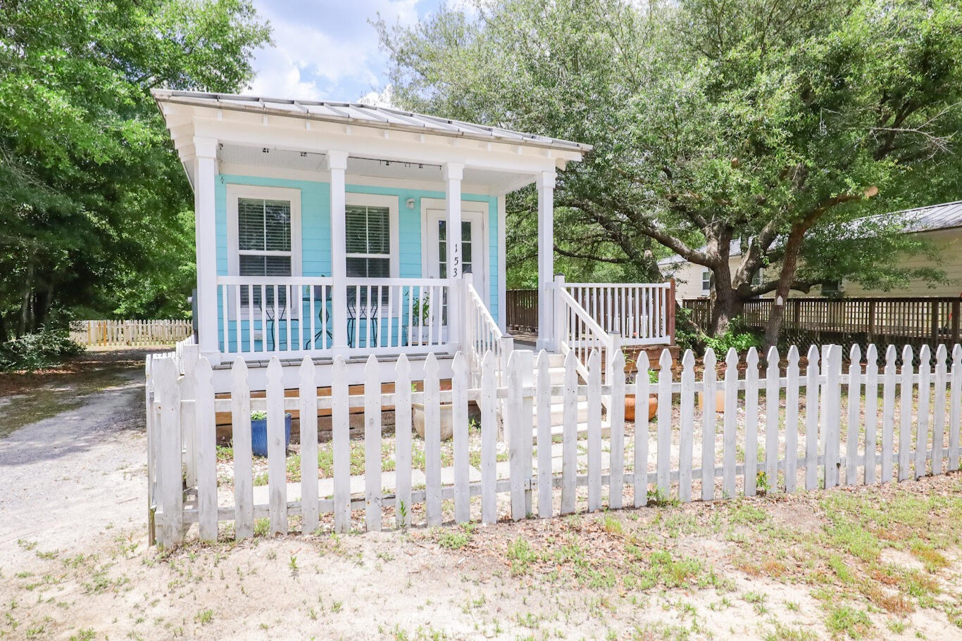 Turquoise cottage with a covered porch behind a white picket fence, surrounded by mature trees.