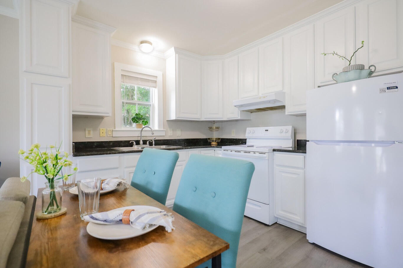 White kitchen with dark countertops, a window over the sink, and a dining table with turquoise chairs.