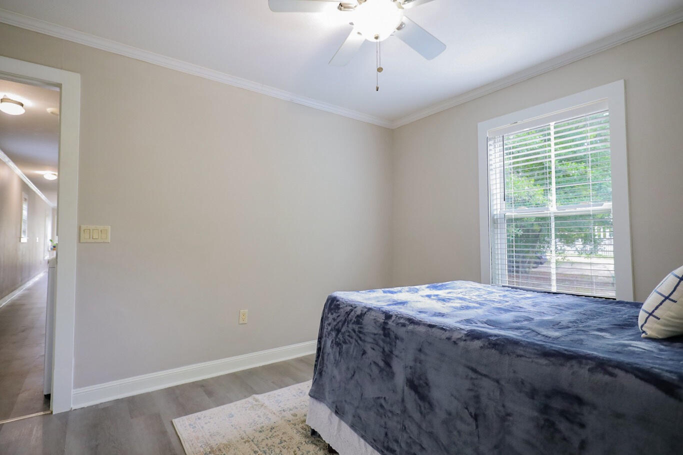 Bedroom with a blue-gray bedspread, large window with blinds, and ceiling fan.