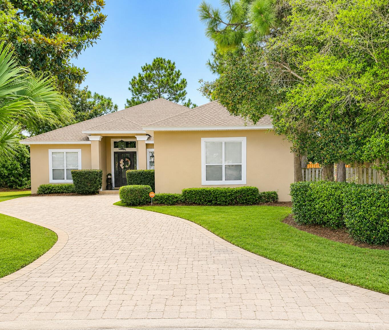 Single-story beige home with a curved paver driveway, trimmed hedges, and mature trees.