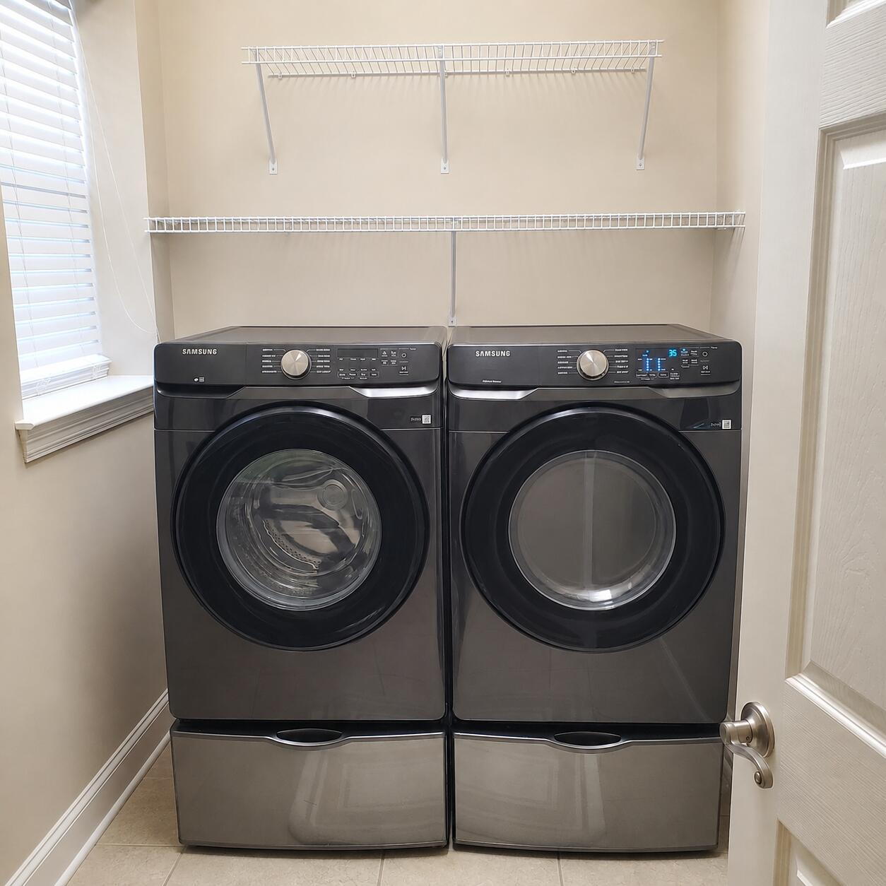 Laundry room with matching Samsung front-loading washer and dryer beneath white wire shelves.