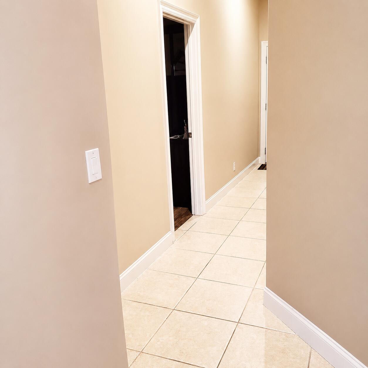 Tiled hallway with beige walls and an open doorway to a darker room.