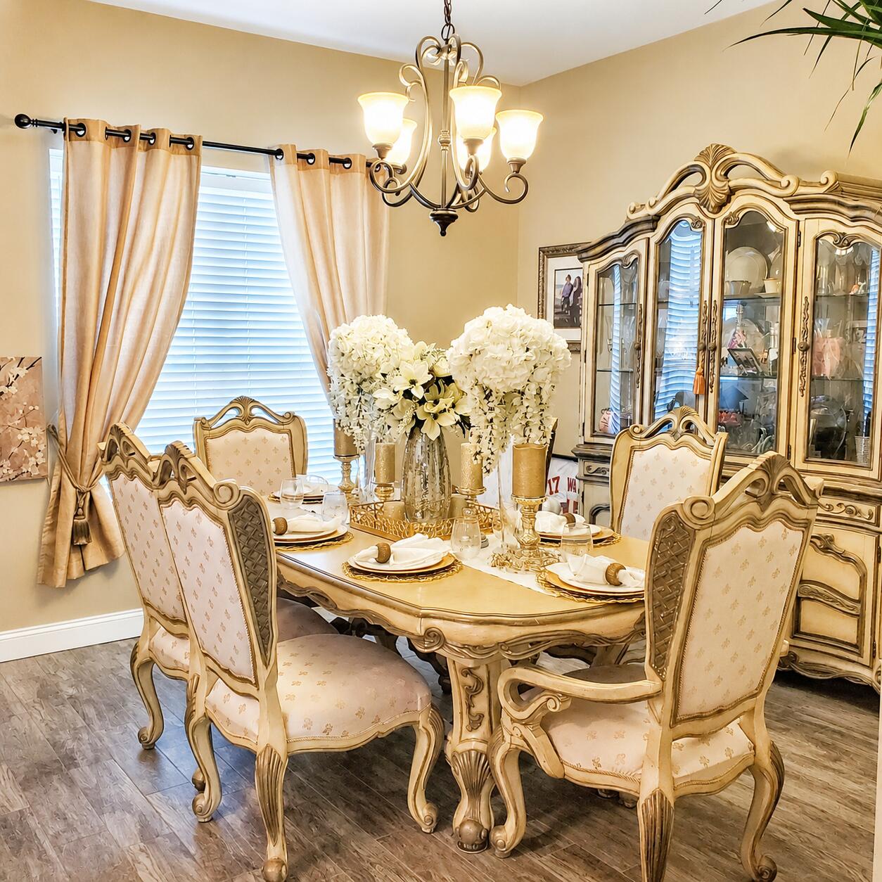 Ornate cream-and-gold dining set beneath a chandelier, with a glass-front china cabinet and flower centerpiece.
