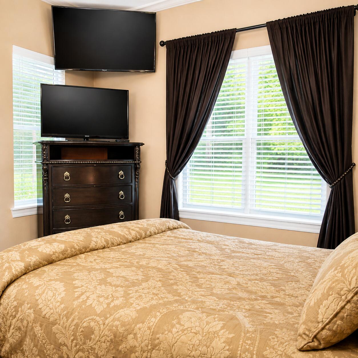 Bedroom with a floral bed, two televisions, and dark curtains framing bright windows.