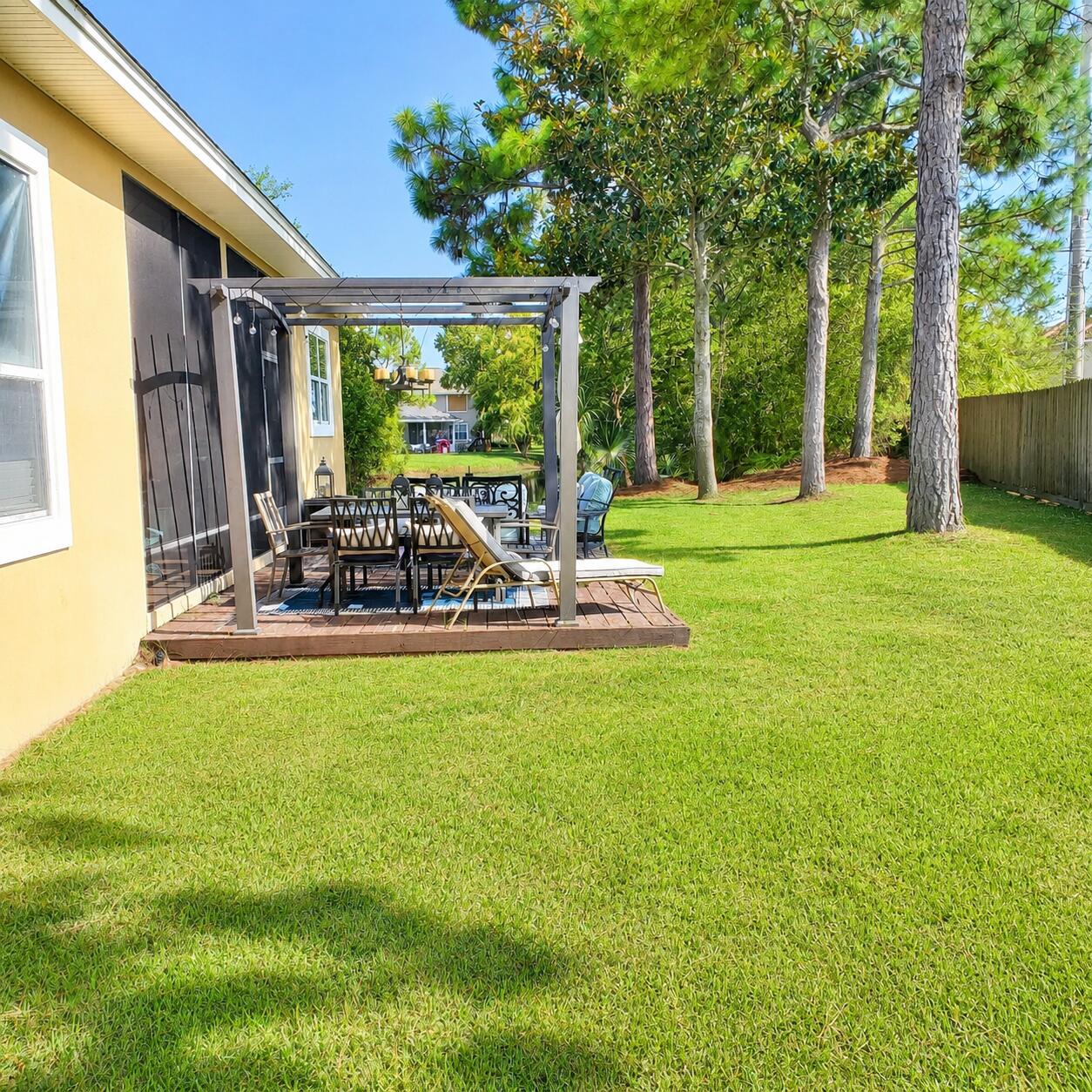 Backyard patio with outdoor dining furniture beneath a pergola beside a spacious lawn and mature trees.