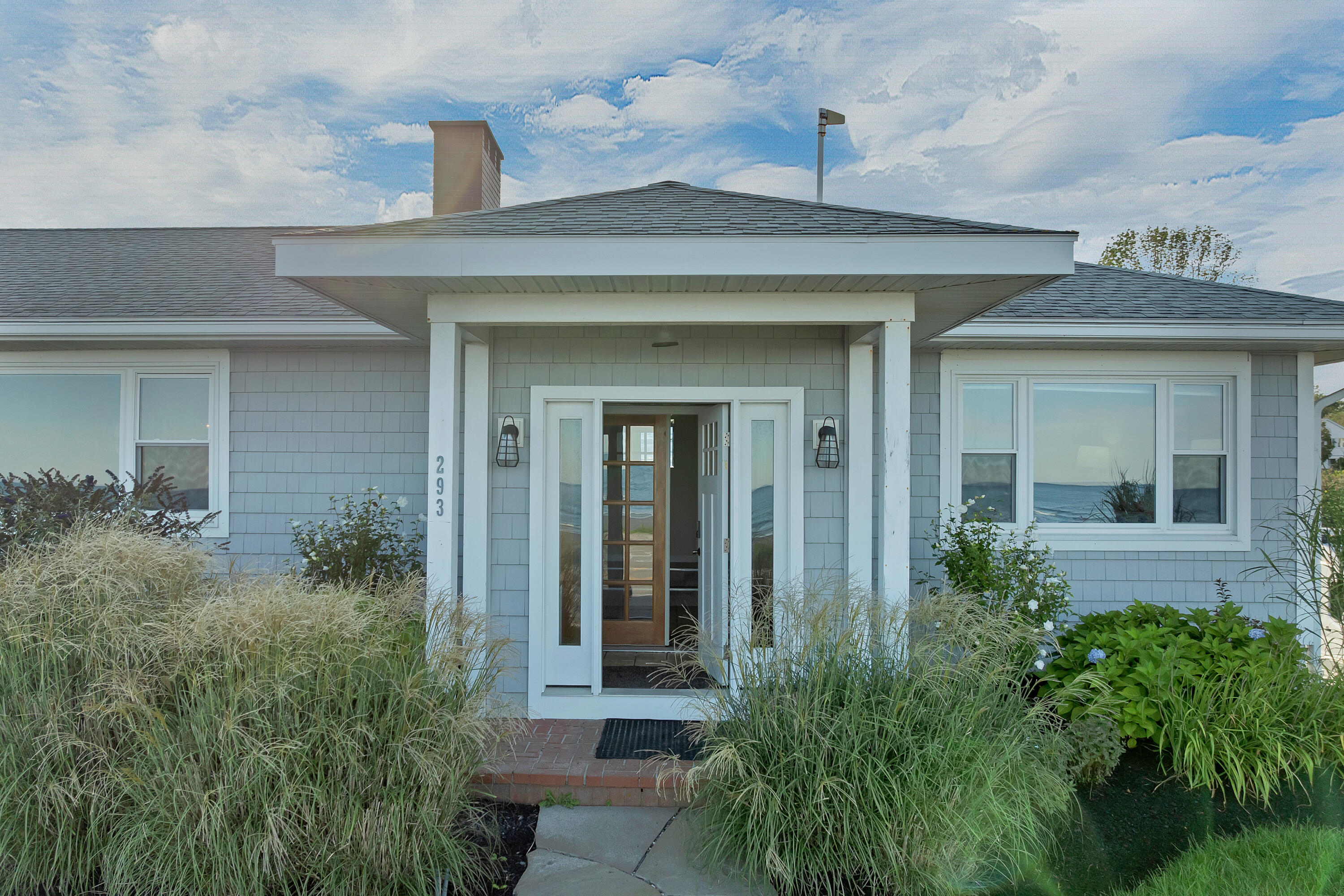 Gray-shingled coastal house with a covered entry, ornamental grasses, and ocean visible through the windows.
