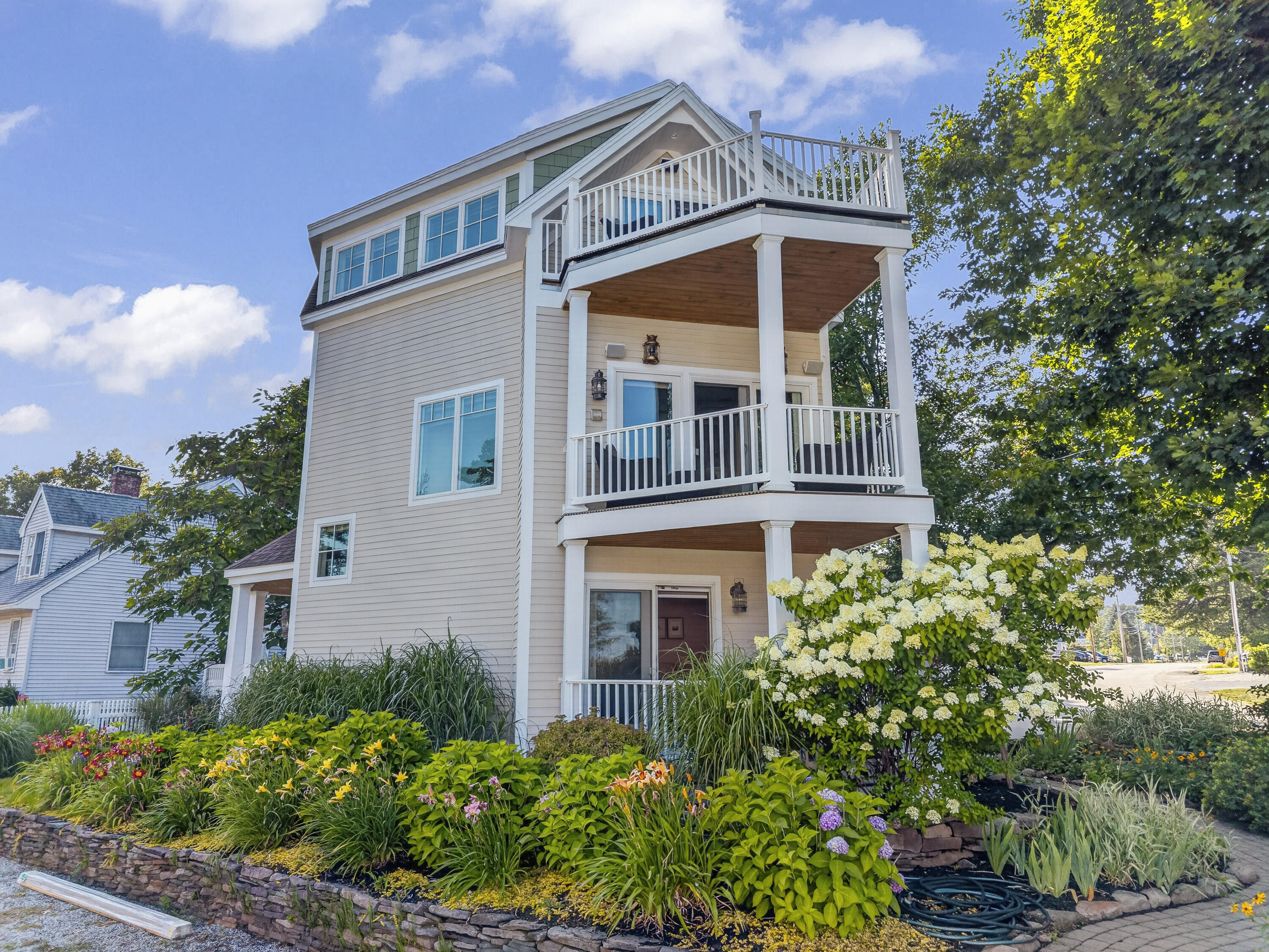 Light-colored multi-story home with two balconies, framed by lush flowering gardens.