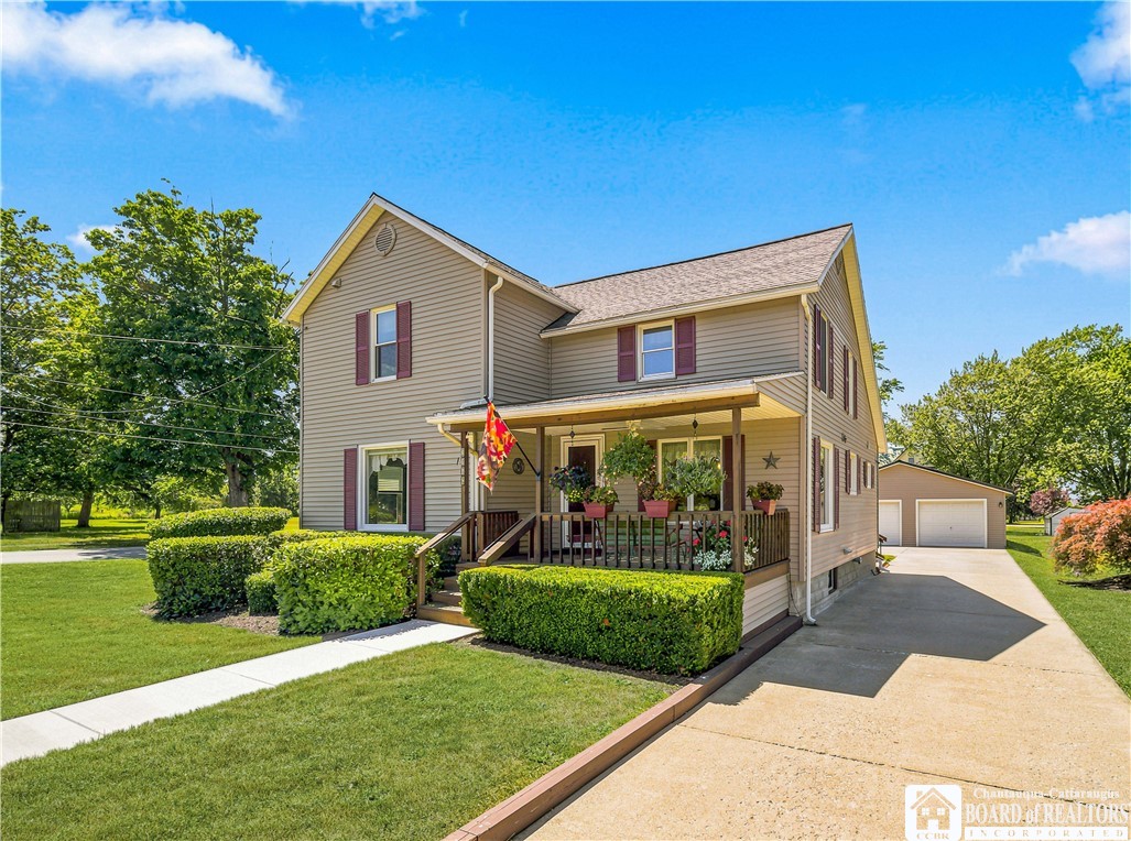 Two-story beige house with burgundy shutters, a plant-filled covered porch, trimmed hedges, and a driveway to a detached garage.