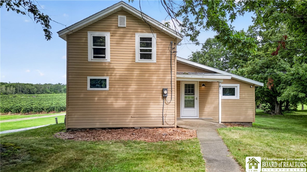 Tan two-story house with white trim, a front walkway, and a lawn bordered by trees and fields.