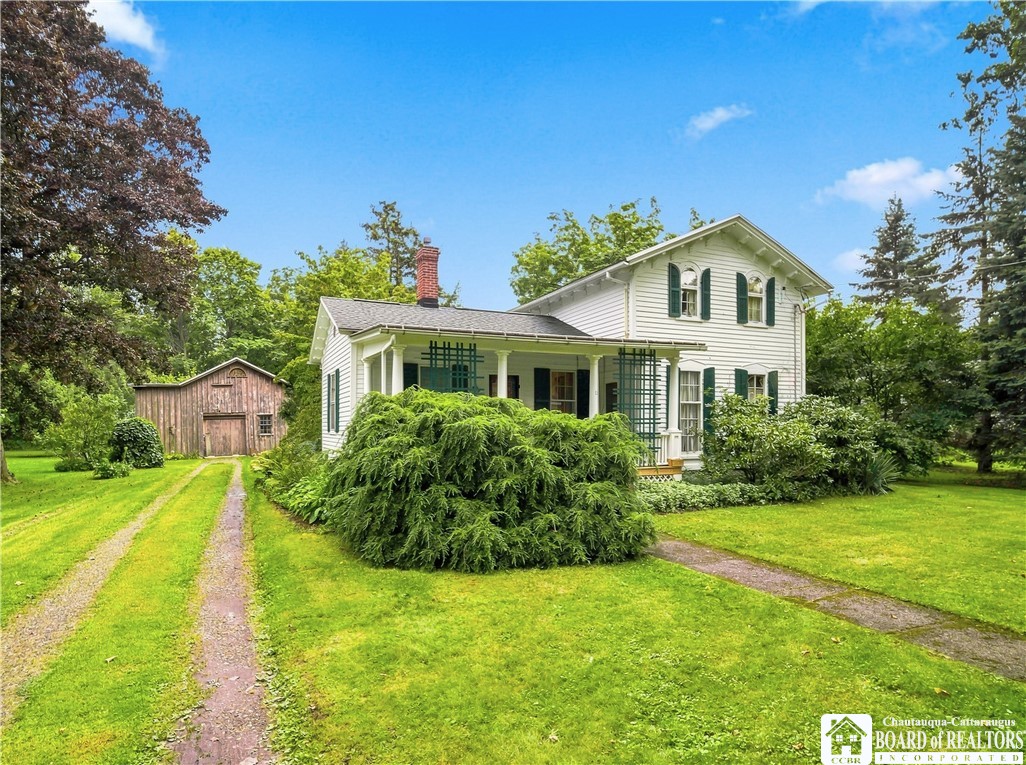 White two-story farmhouse with green shutters, a covered porch, and a small wooden barn beside the driveway.