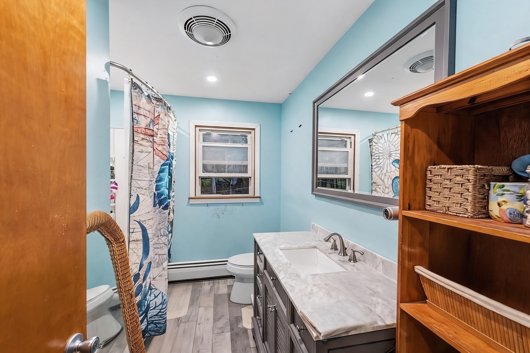 Bathroom with pale blue walls, marble-look vanity, large mirror, and patterned shower curtain.