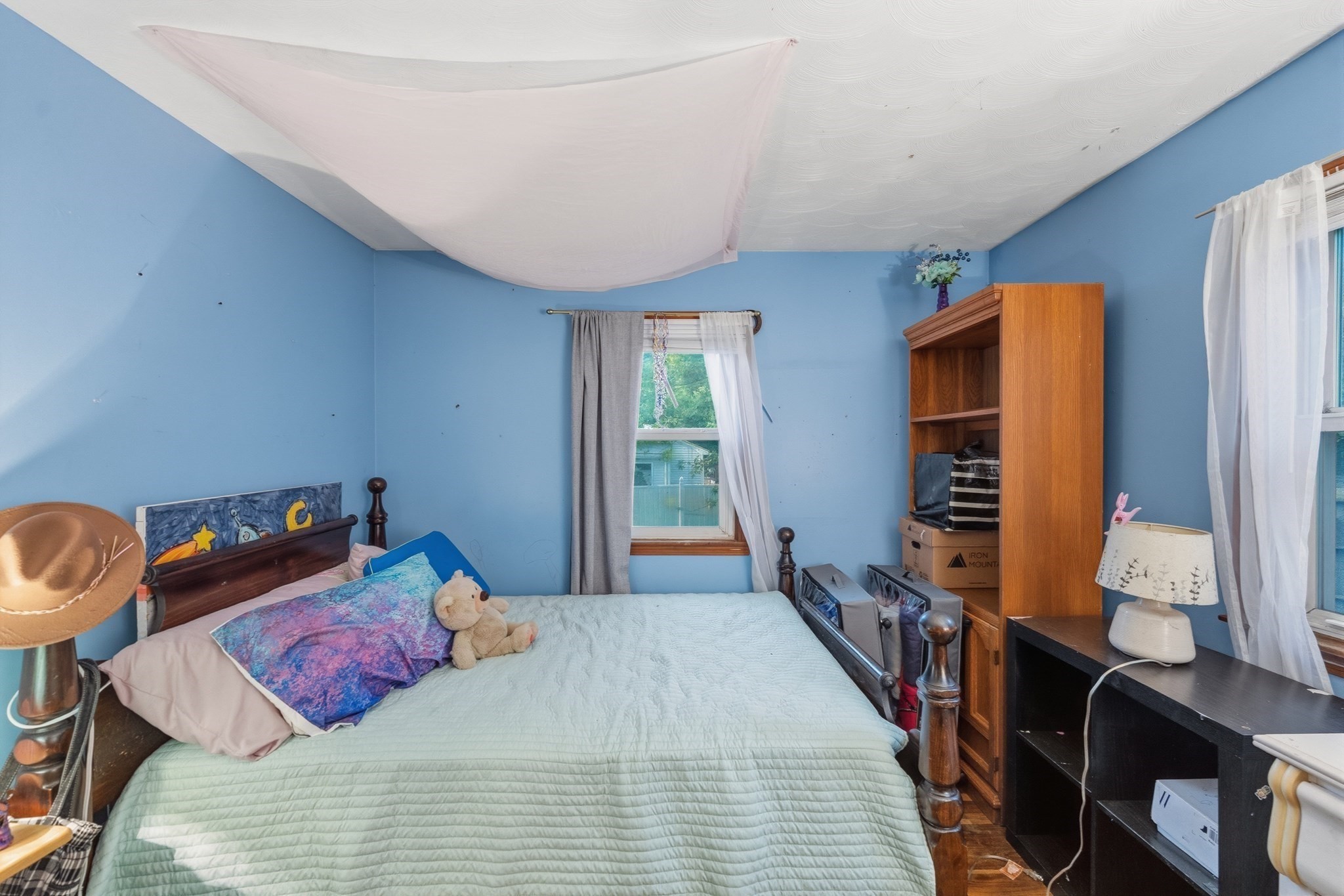 Bedroom with blue walls, a wooden bed, two curtained windows, and a tall bookcase.