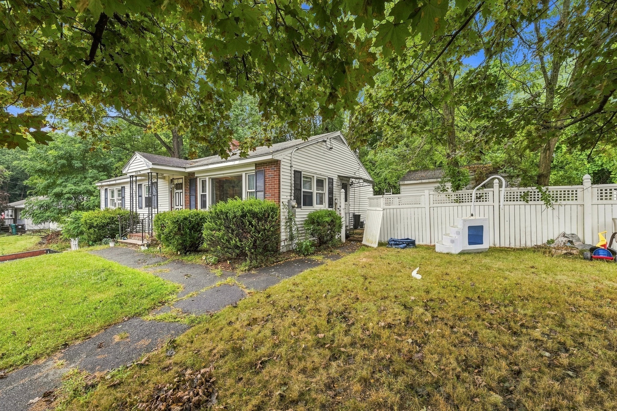 Single-story house with a covered porch, brick facade, leafy yard, and white privacy fence.