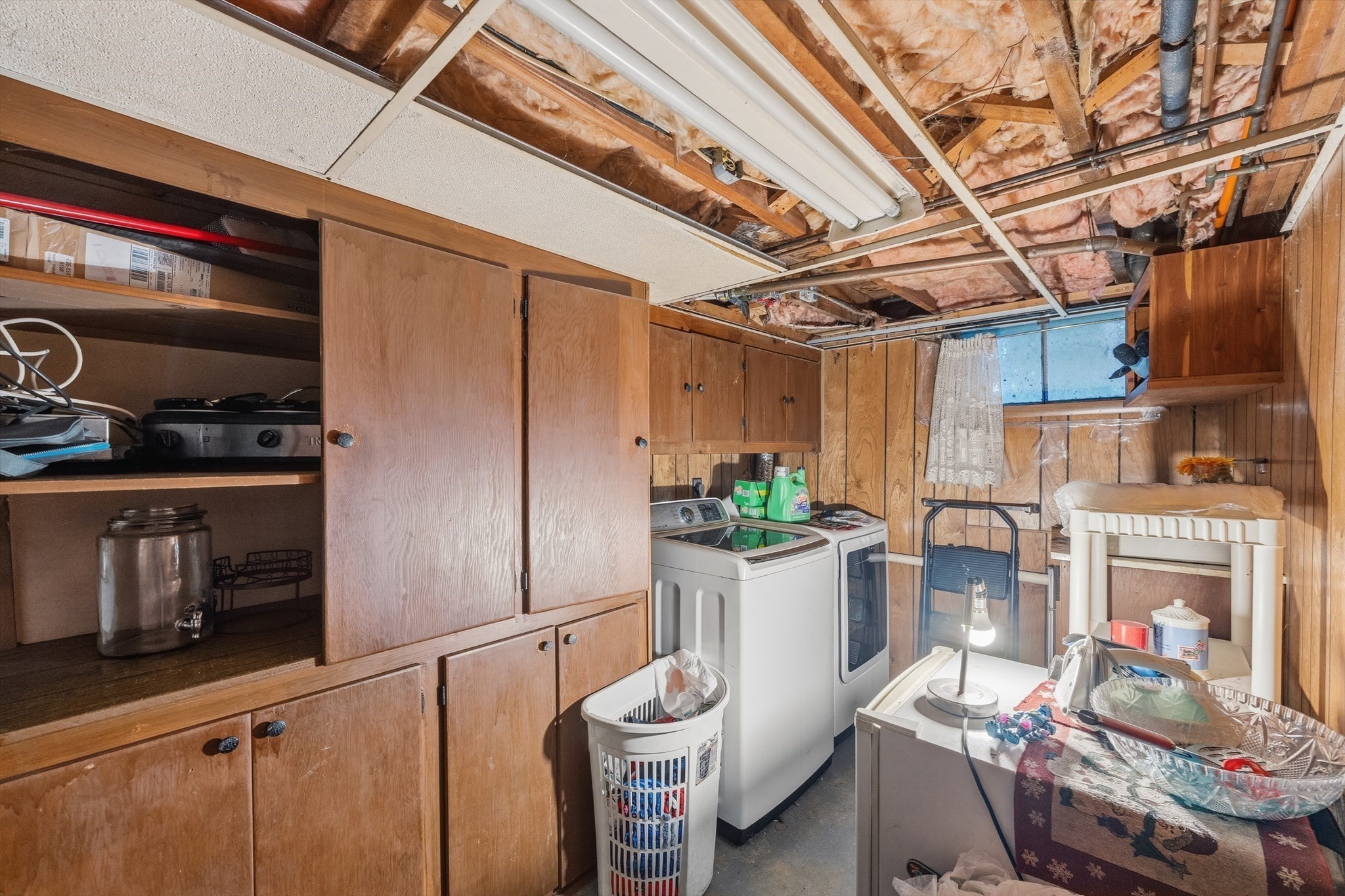 Basement laundry room with wooden storage cabinets, washer and dryer, and exposed ceiling insulation.