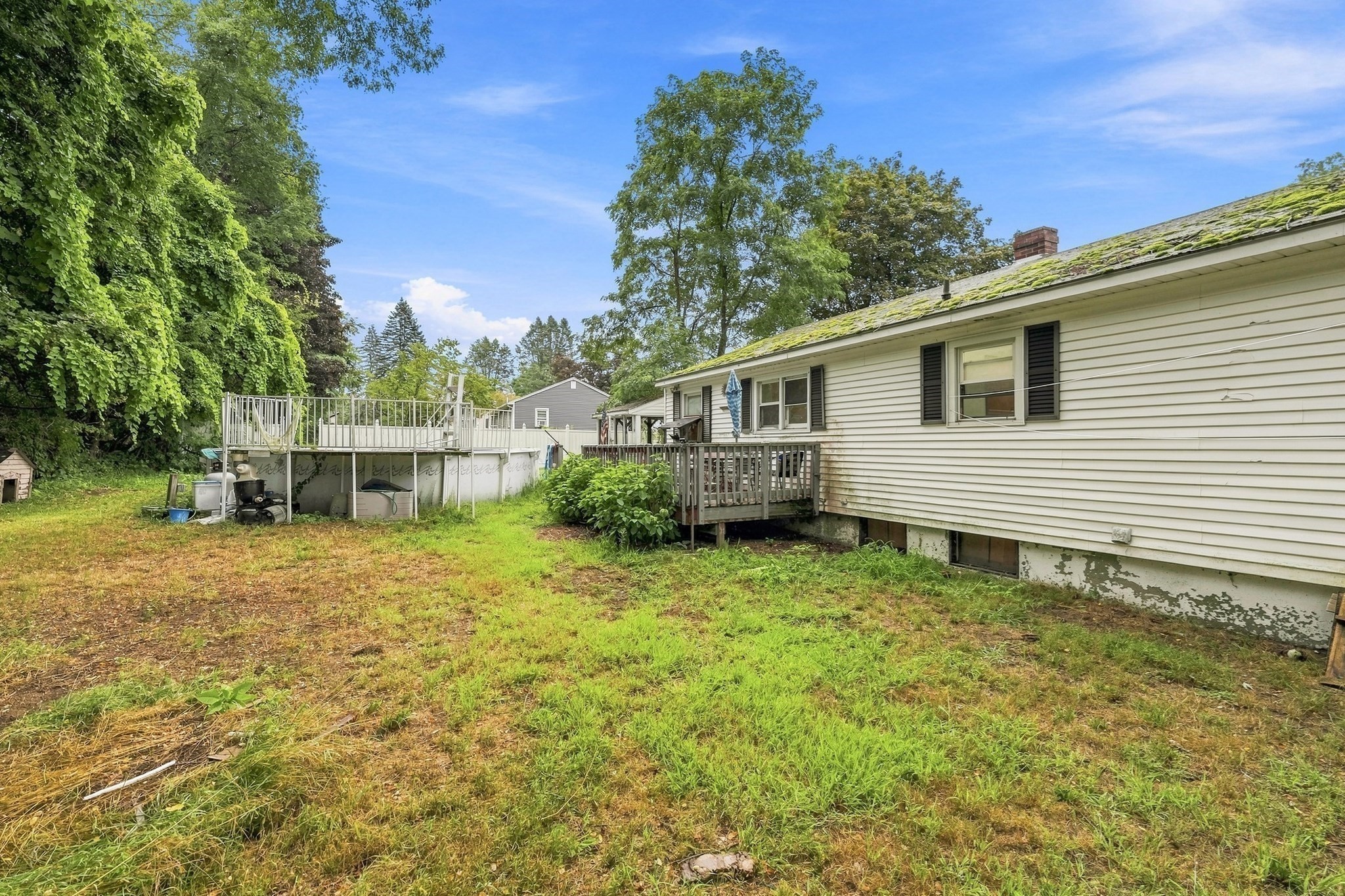 White-sided home with a small deck and raised platform beside a grassy, tree-lined yard.