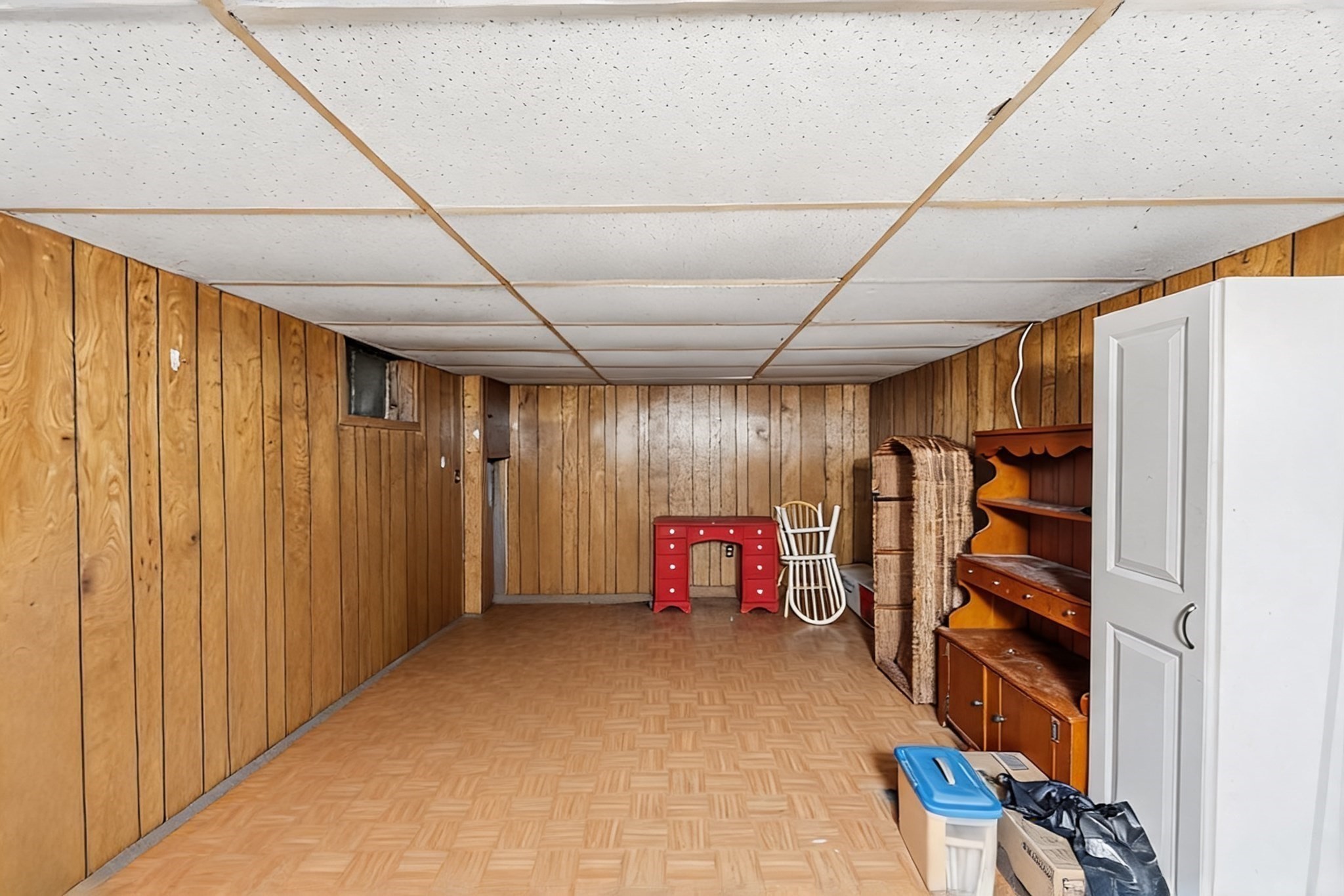 Basement room with wood-paneled walls, patterned flooring, and stored furniture along one side.